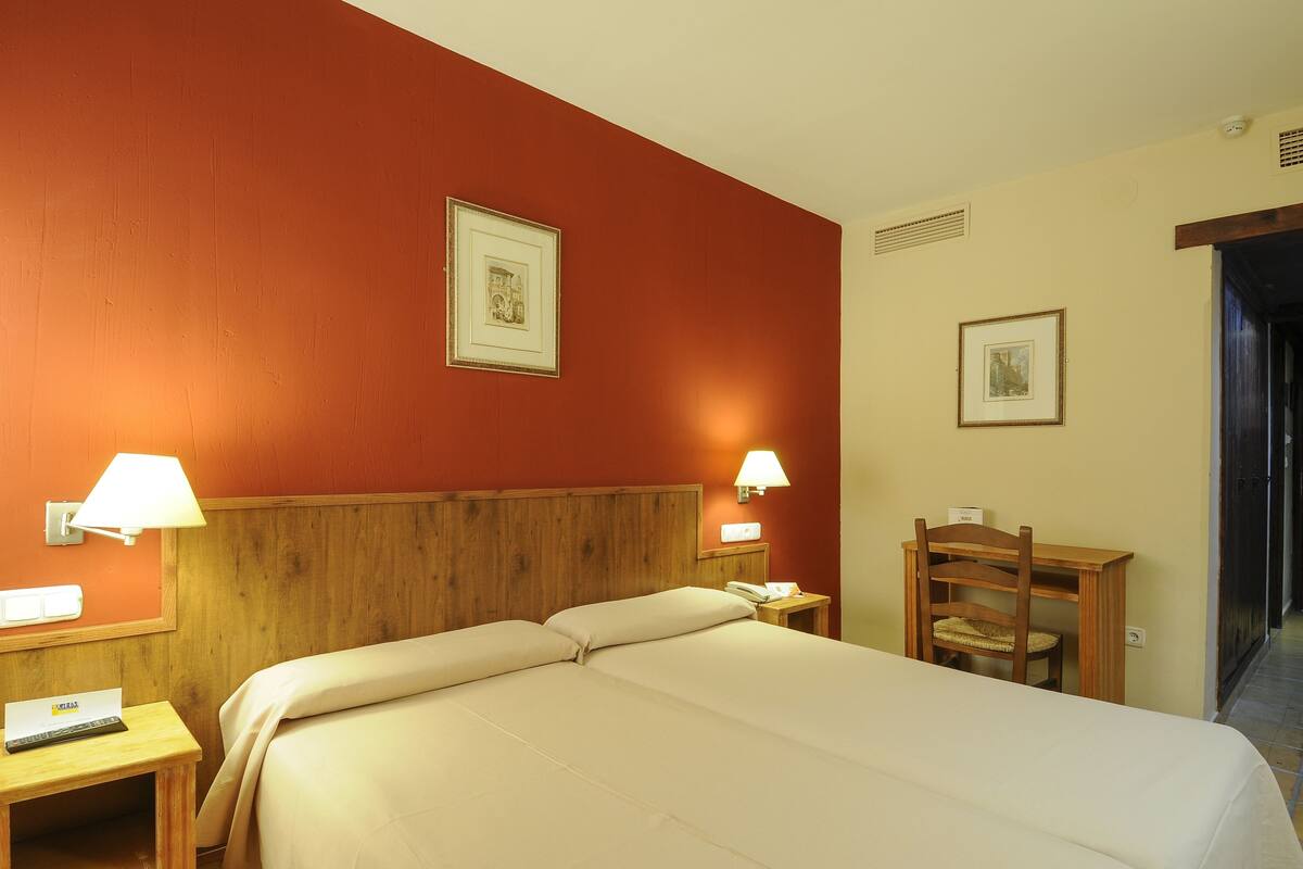 Un cuarto de hotel con una cama grande, dos lámparas de noche, un cabecero de madera, un escritorio con una silla y dos cuadros enmarcados en las paredes.