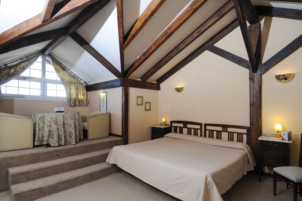 Un dormitorio con una cama grande, una mesita con mantel decorativo, una silla y una mesita de noche con lámpara.