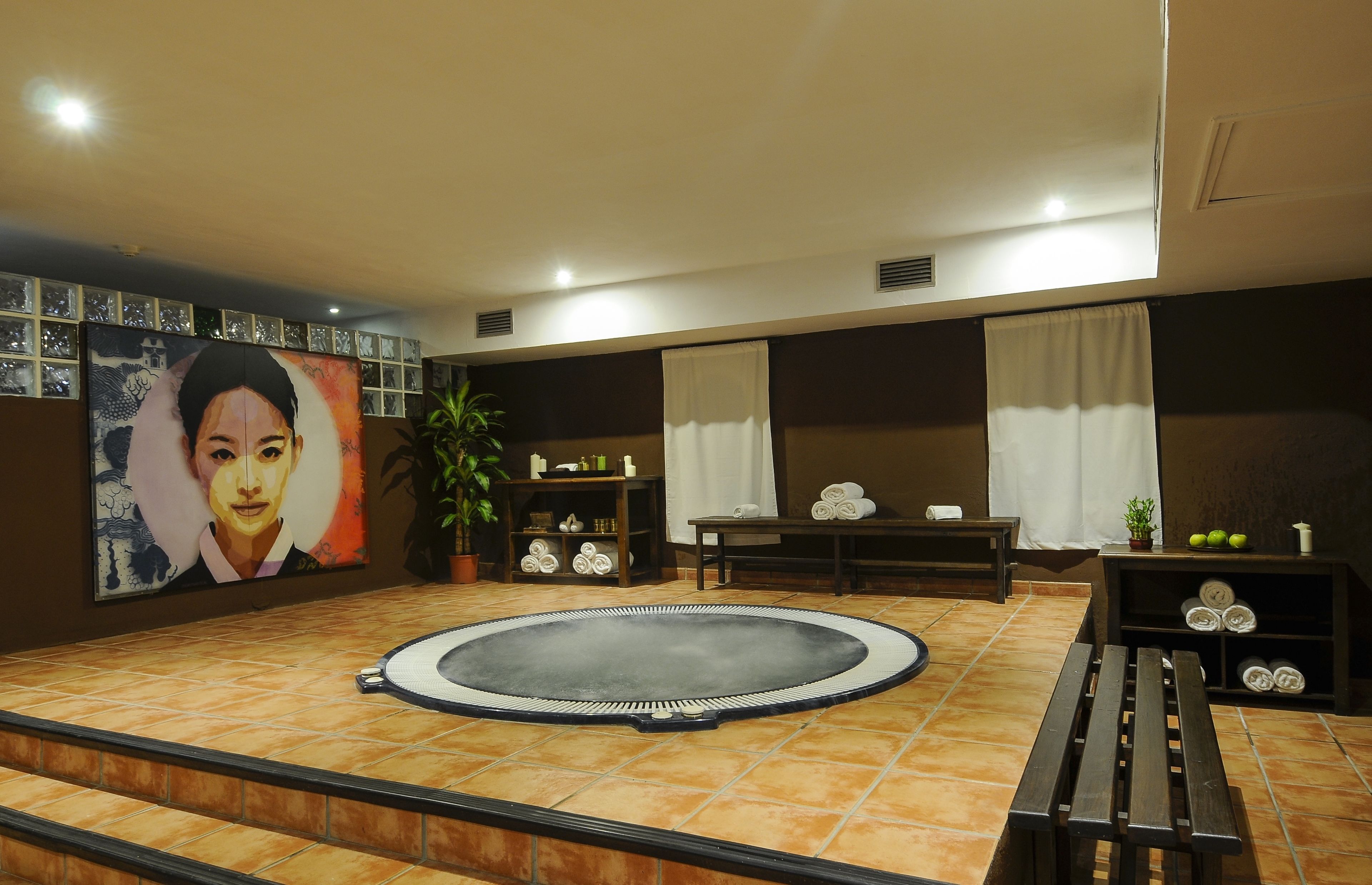 A room with a circular hot tub, a large painting, a potted plant, a bench, and a shelf with towels and toiletries.