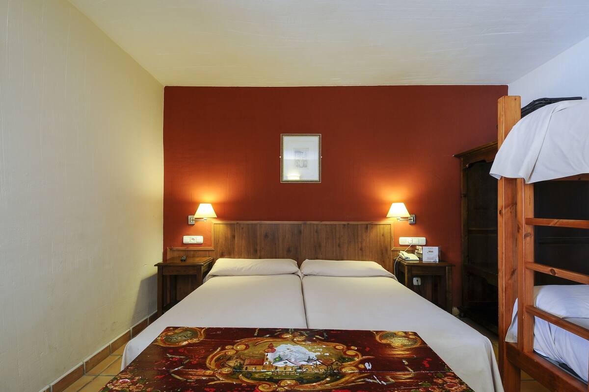 Una cama doble con cabecera decorativa, dos lámparas de noche, una mesita de noche pequeña y una cama litera de madera a la derecha.