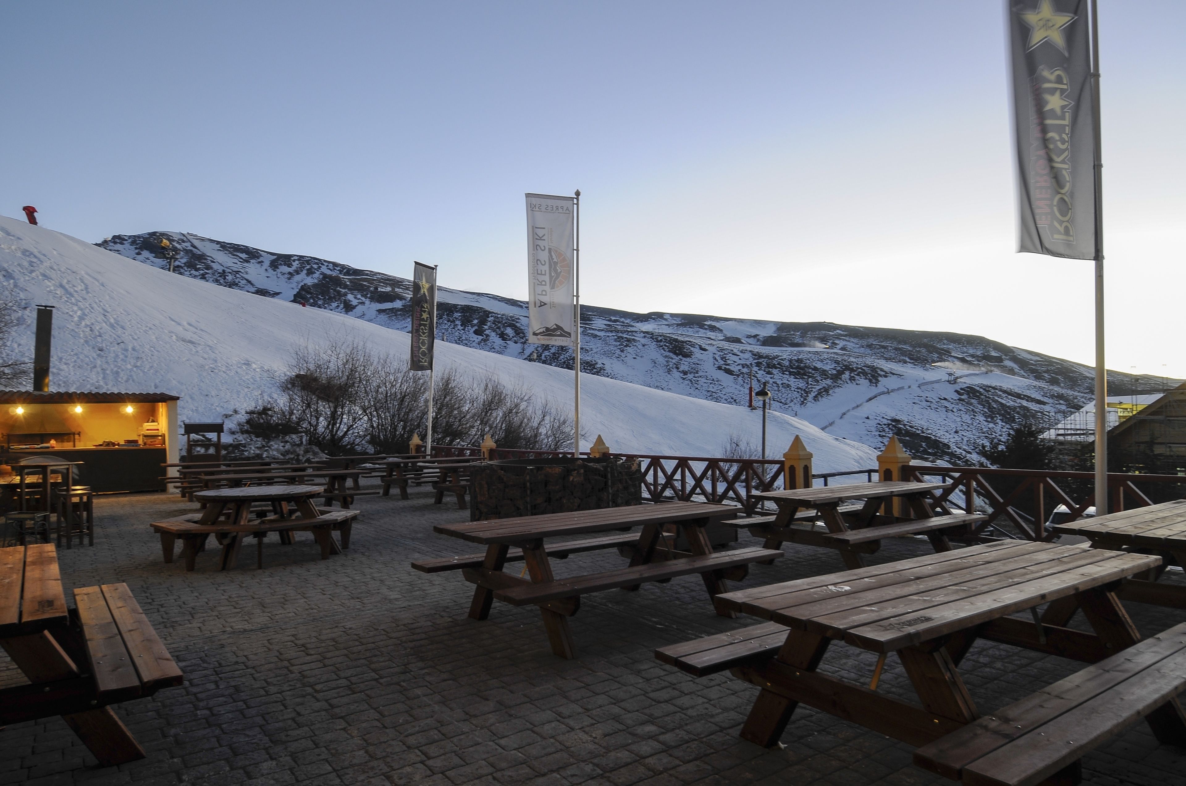 A snow-covered outdoor area with picnic tables and a building with a bar.