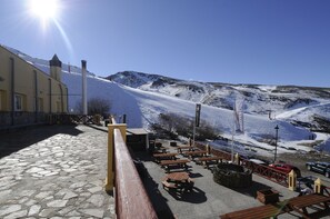 A snow-covered outdoor seating area with stone flooring, wooden tables, and benches, overlooking a mountainous landscape.