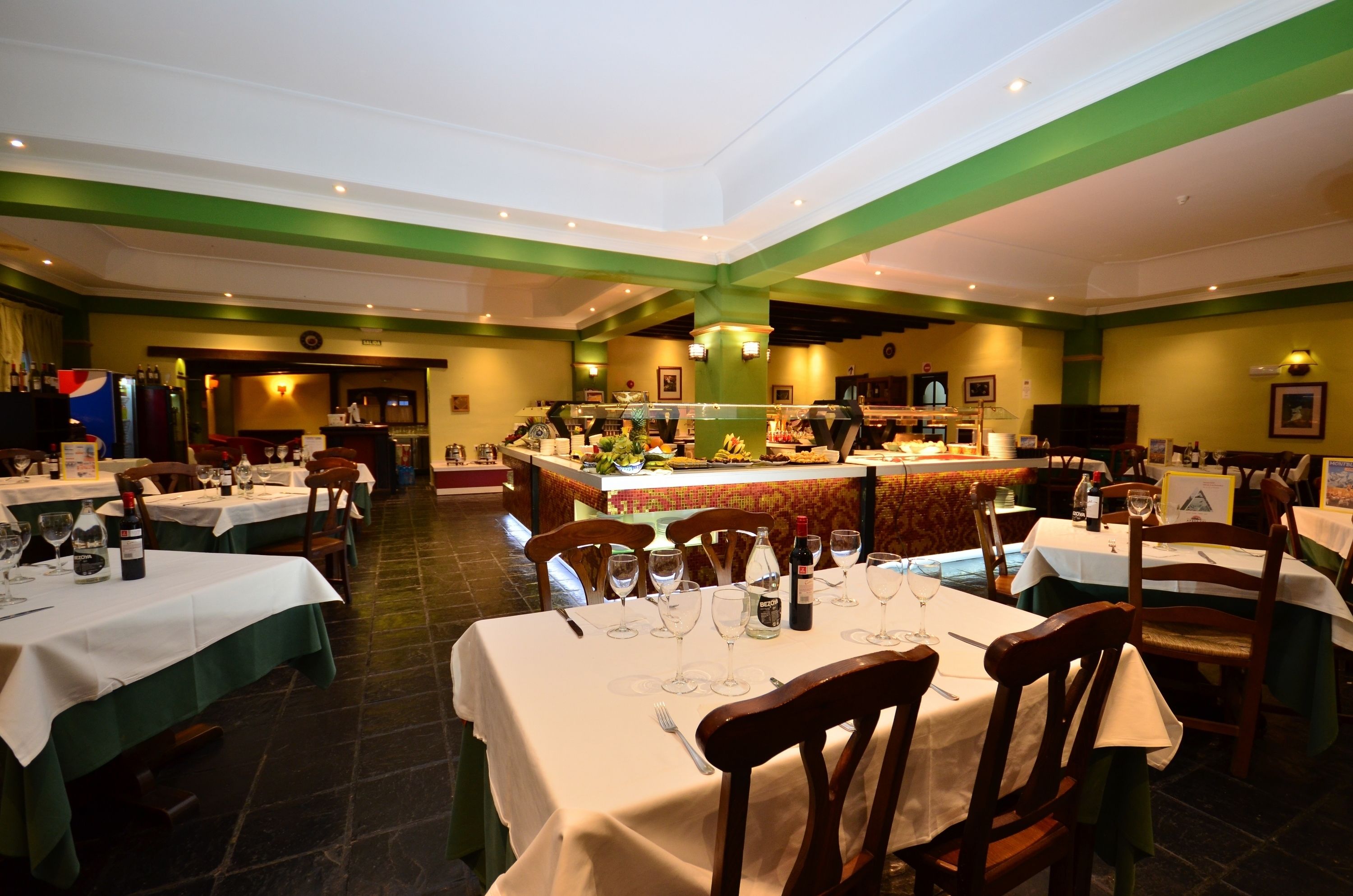 Un restaurante con varias mesas, cada una dispuesta con copas de vino y cubiertos, y una zona de bar central con una variedad de alimentos y bebidas.