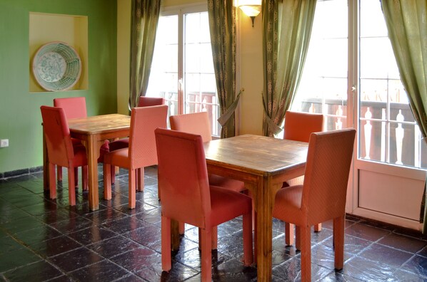 Uma sala de jantar com mesa de madeira e seis cadeiras vermelhas, parede verde e vista através de uma porta de vidro.