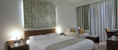 A hotel room with a large bed, a desk, a chair, a nightstand, and a wall with a floral pattern.