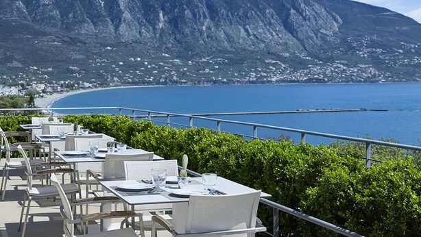 A seaside restaurant with outdoor seating overlooking a beach and mountains. - Pharae Palace Hotel (Kalamata)