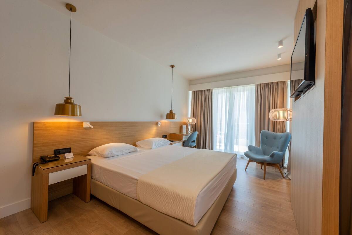 Ein Hotelzimmer mit einem großen Bett, Nachttischen, einem Schreibtisch, einem Sessel und einem an der Wand befestigten Fernseher.