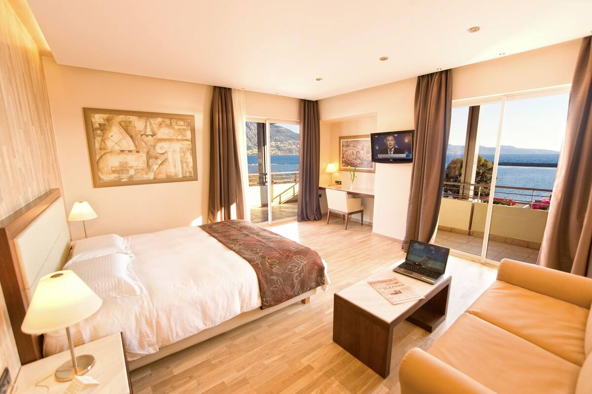 Ein Hotelzimmer mit Bett, Schreibtisch, Laptop, Sofa und einem Balkon mit Meerblick.