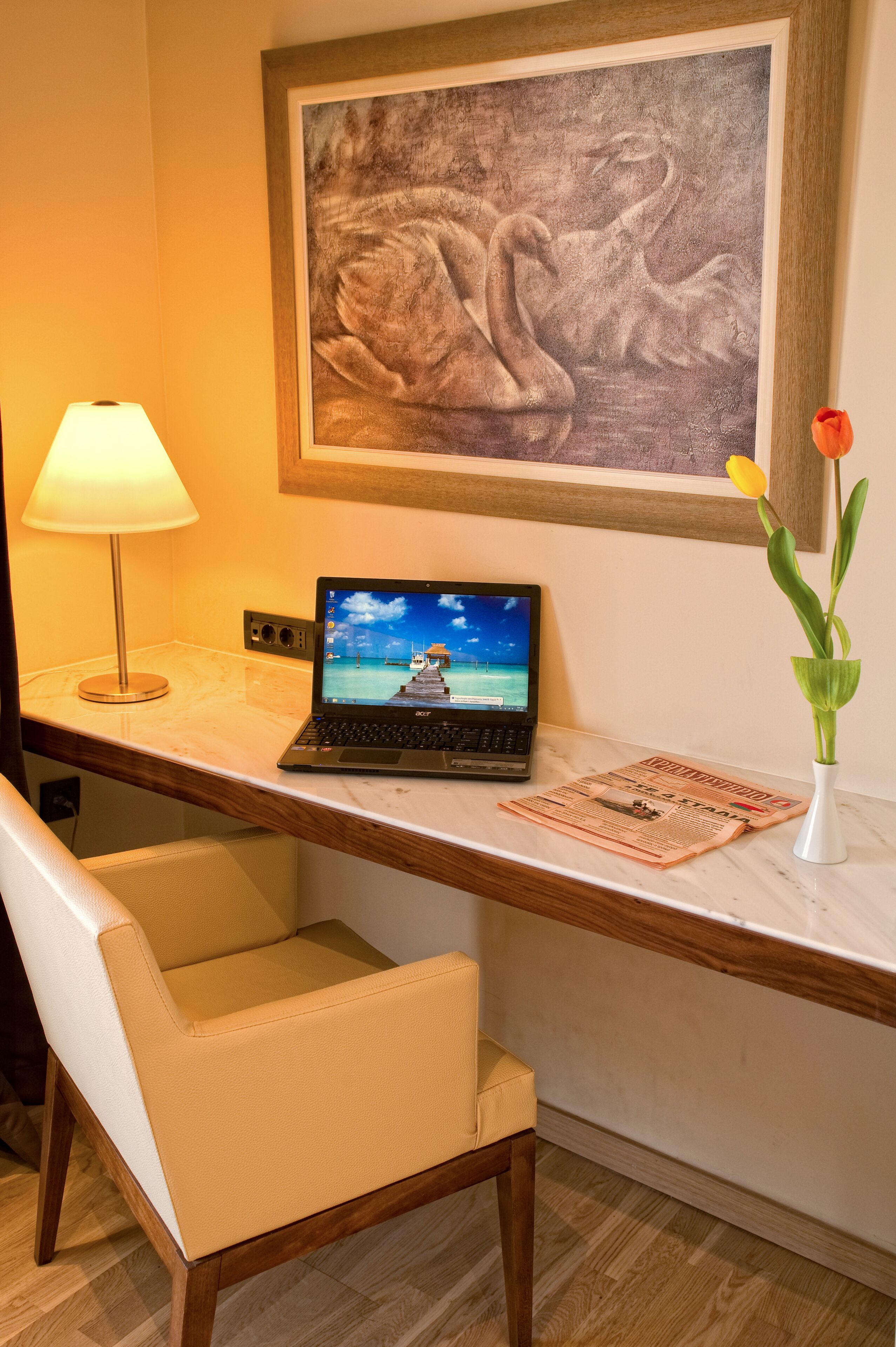 Habitación de hotel con escritorio, computadora portátil, lámpara, silla, un cuadro y un florero con una flor.