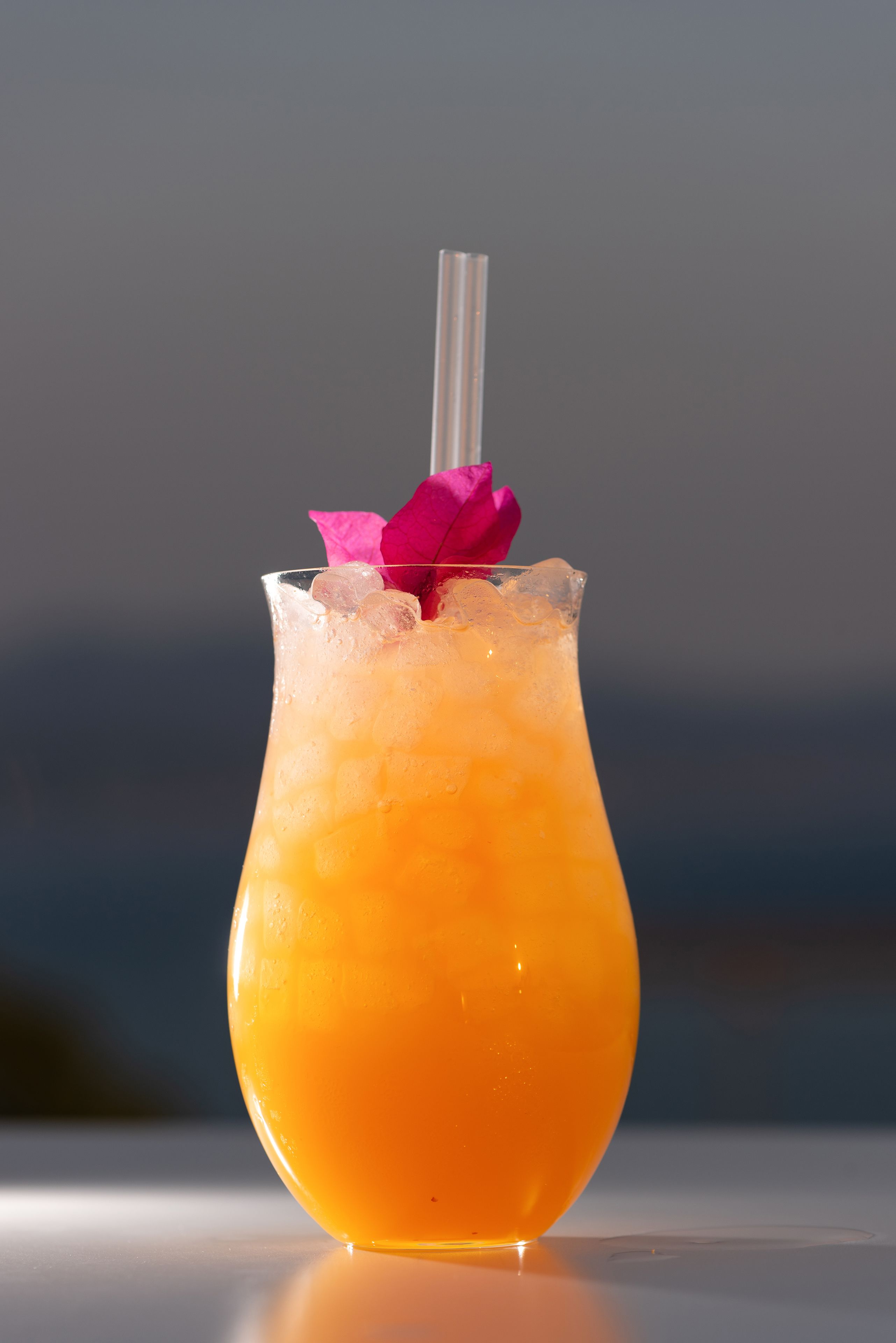 A cocktail in a glass with a pink flower garnish and a straw.
