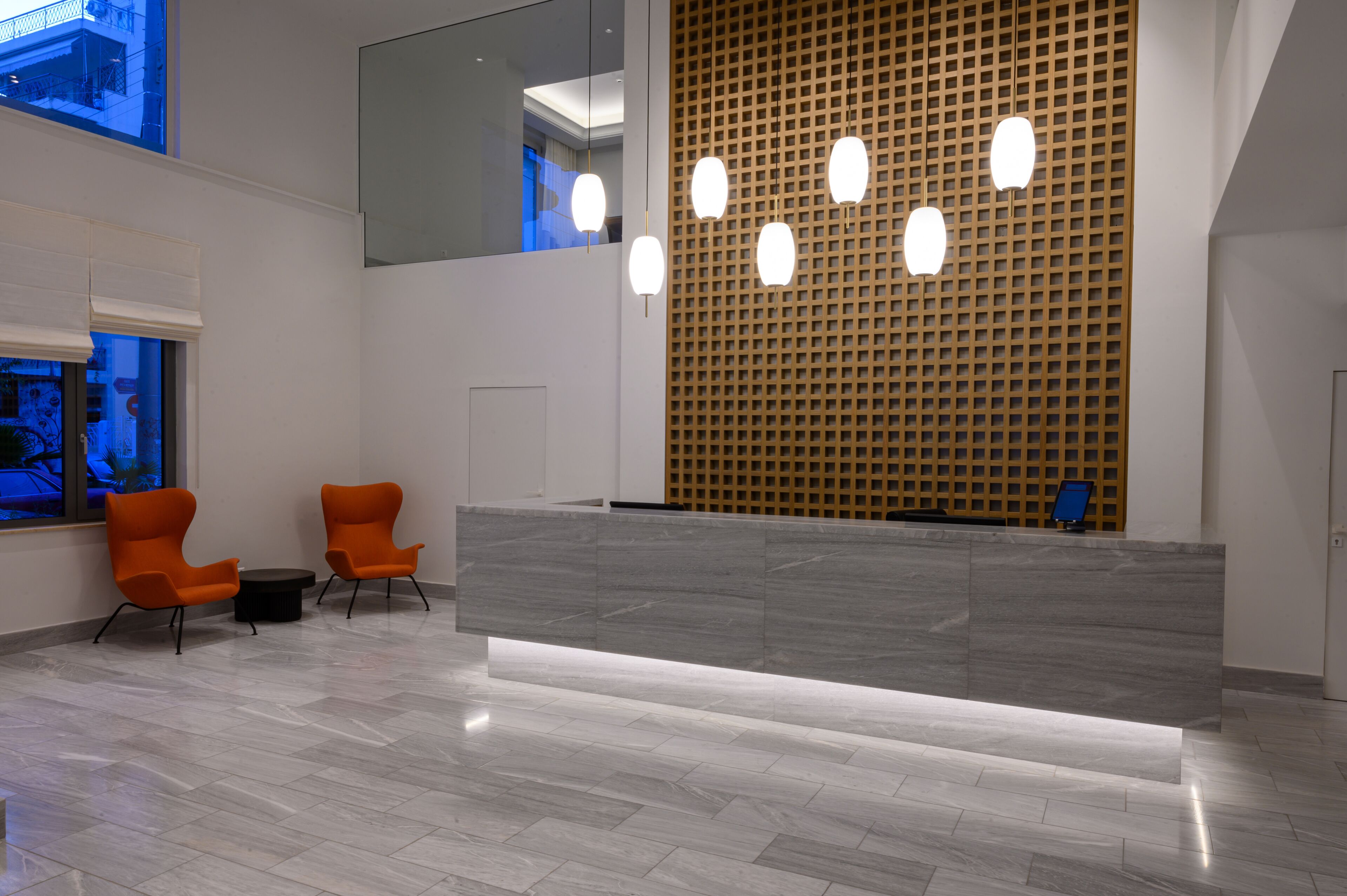 Un área de recepción con mostrador de mármol, dos sillas color naranja, pared de madera con diseño de cuadrícula y un ventanal grande.