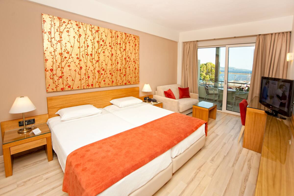 Ein Hotelzimmer mit einem großen Bett, einer Sitzecke, einem Fernseher, einem Balkon mit Aussicht und einem Bild an der Wand.