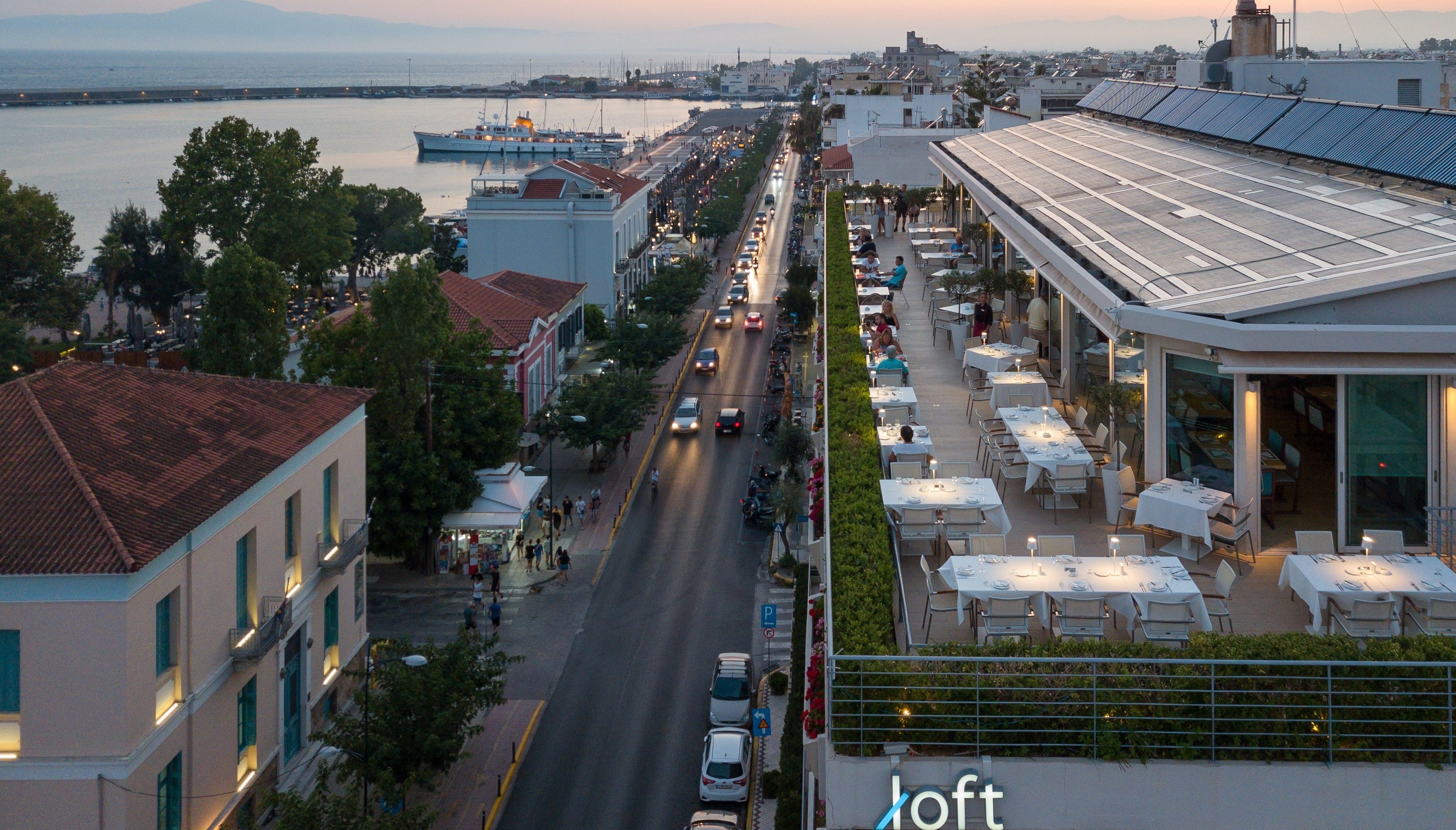 Uma rua costeira com áreas de refeições ao ar livre, um porto de iates e edifícios com telhados de barro vermelhos.