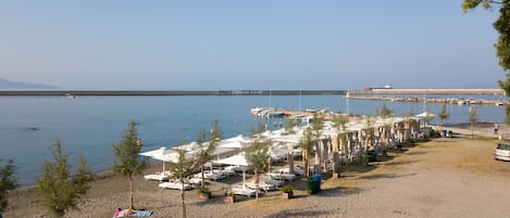Una zona de playa con filas de tumbonas y sombrillas, una playa de arena y un mar tranquilo con barcos a lo lejos.