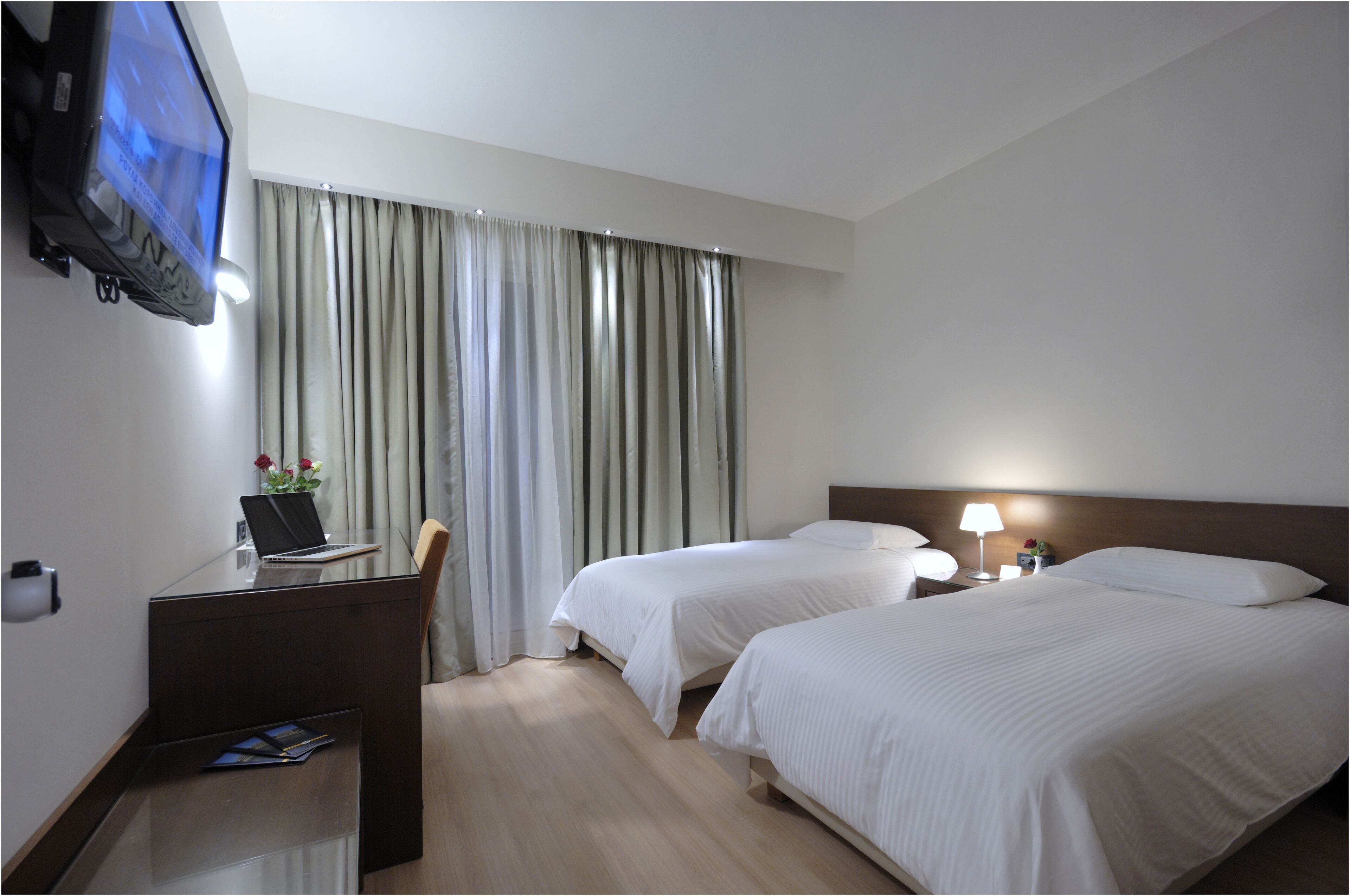 A hotel room with two beds, a desk with a laptop, a TV mounted on the wall, and a window with curtains.