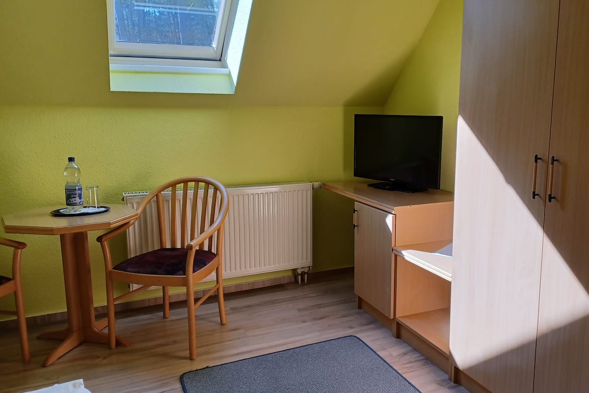 Ein kleiner Raum mit einem Holzbstuhl, einem Tisch mit Flasche und Glas, einem Heizkörper, einem Fernseher auf einem Schrank und einem Schränkchen.