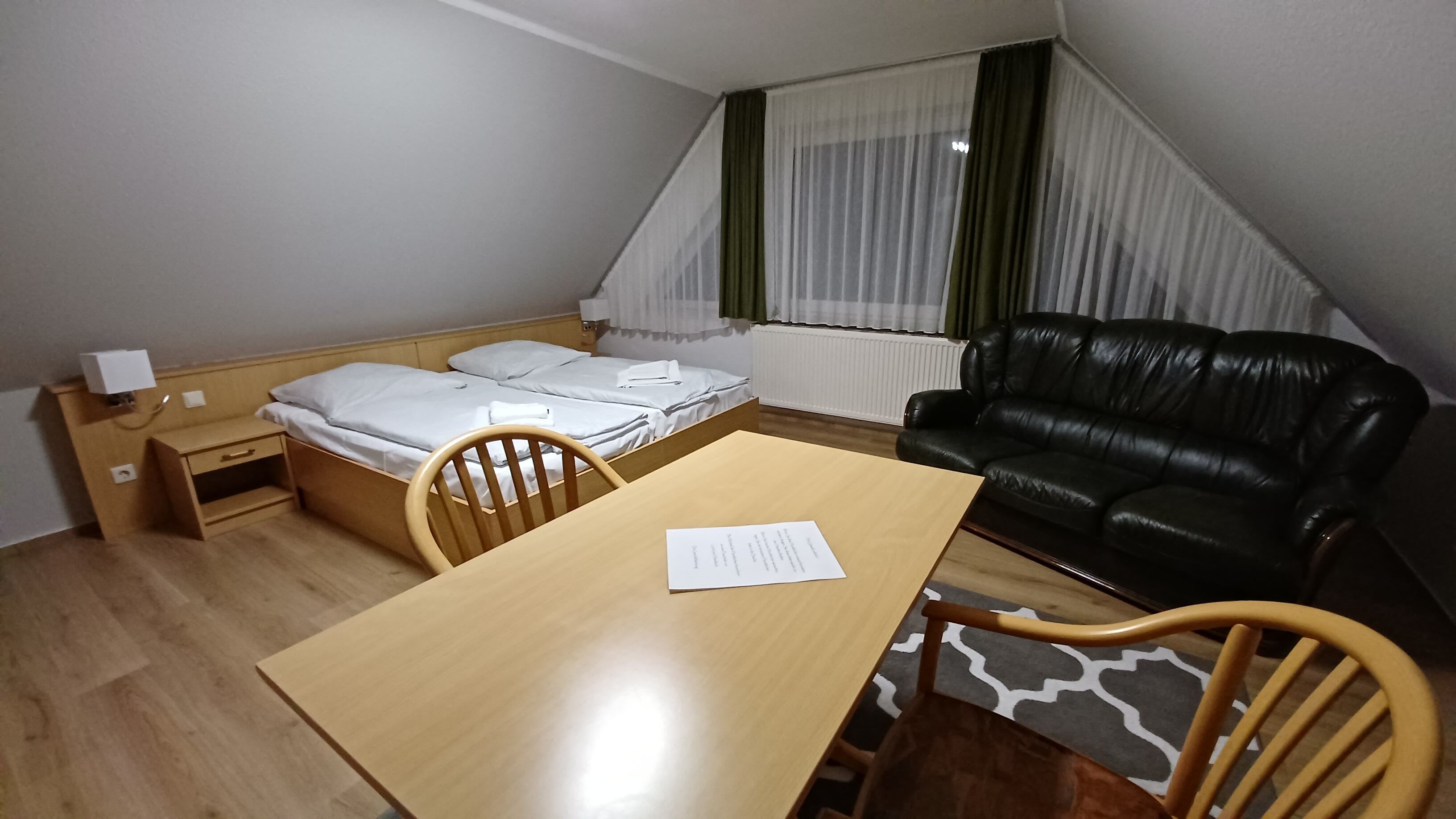 Une petite pièce meublée, comprenant un lit, un bureau en bois, une chaise, un canapé en cuir noir et une fenêtre avec des rideaux.