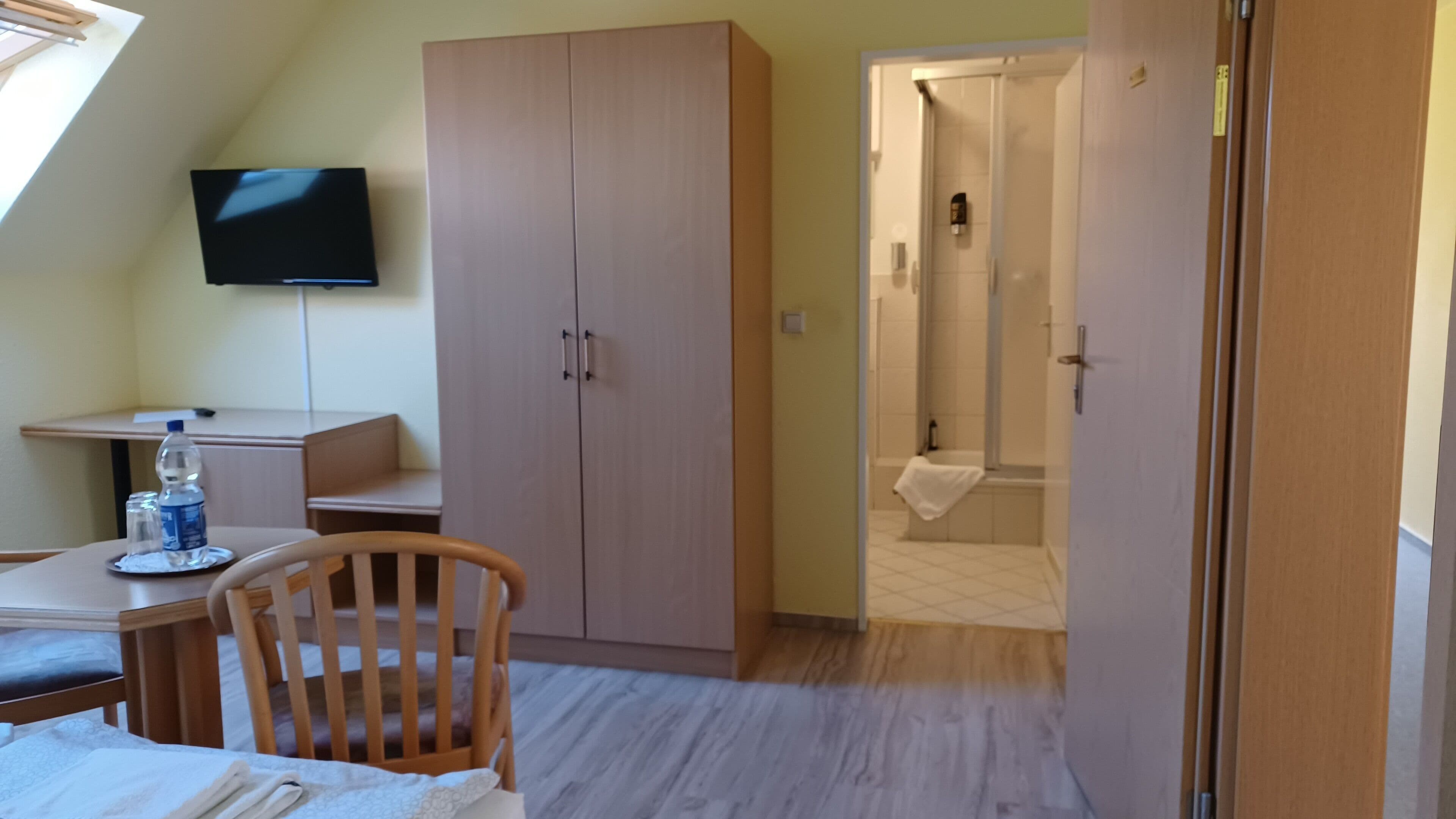 A room with a bed, a desk with a chair, a wardrobe, a television, and a shower area.
