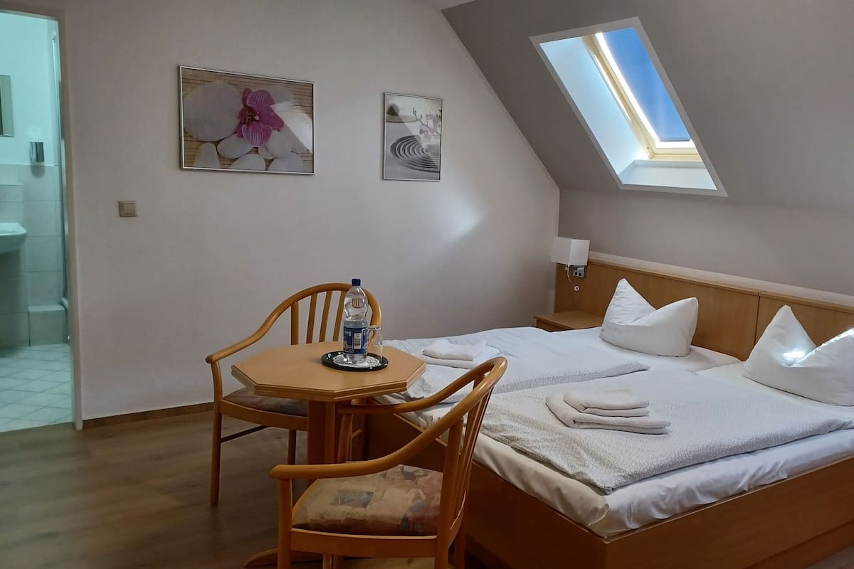 Ein Schlafzimmer mit einem Bett, zwei Kissen, einem Nachttisch, einem Tisch mit Stuhl, einer Wasserflasche und einem Bild an der Wand.