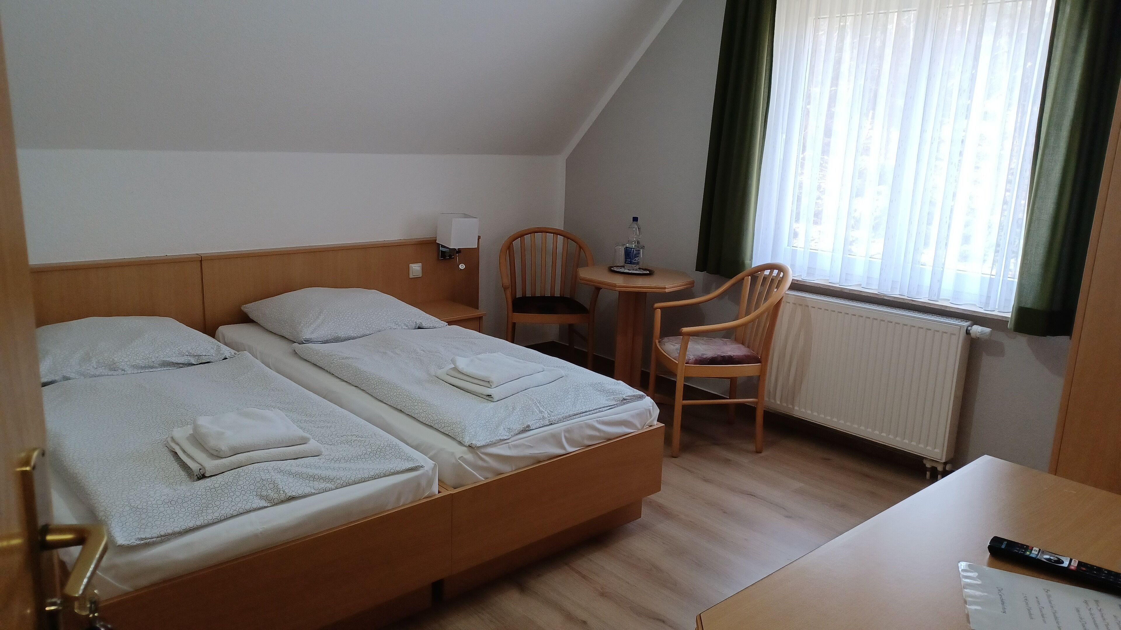 A hotel room with two single beds, a small table with a chair, a desk, and a window with curtains.
