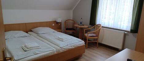 Hotellihuone, jossa on kaksi erillistä sänkyä, pieni pöytä ja tuoli, työpöytä sekä ikkuna, jossa on verhot.