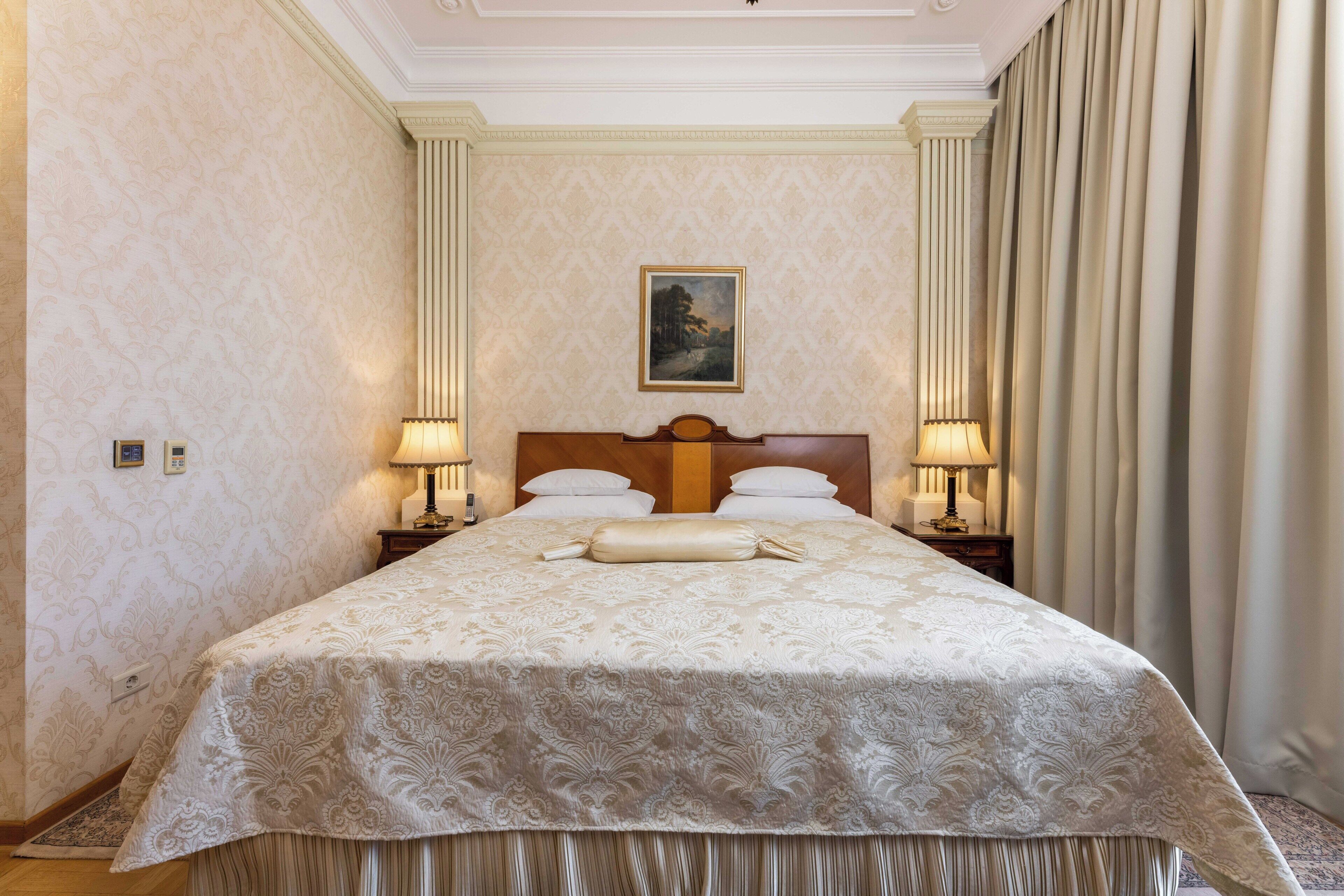 Habitación de hotel con una cama grande, dos lámparas de noche, cabecero con un cuadro y vista al exterior a través de cortinas.