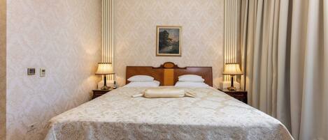 Habitación de hotel con una cama grande, dos lámparas de noche, cabecero con un cuadro y vista al exterior a través de cortinas.