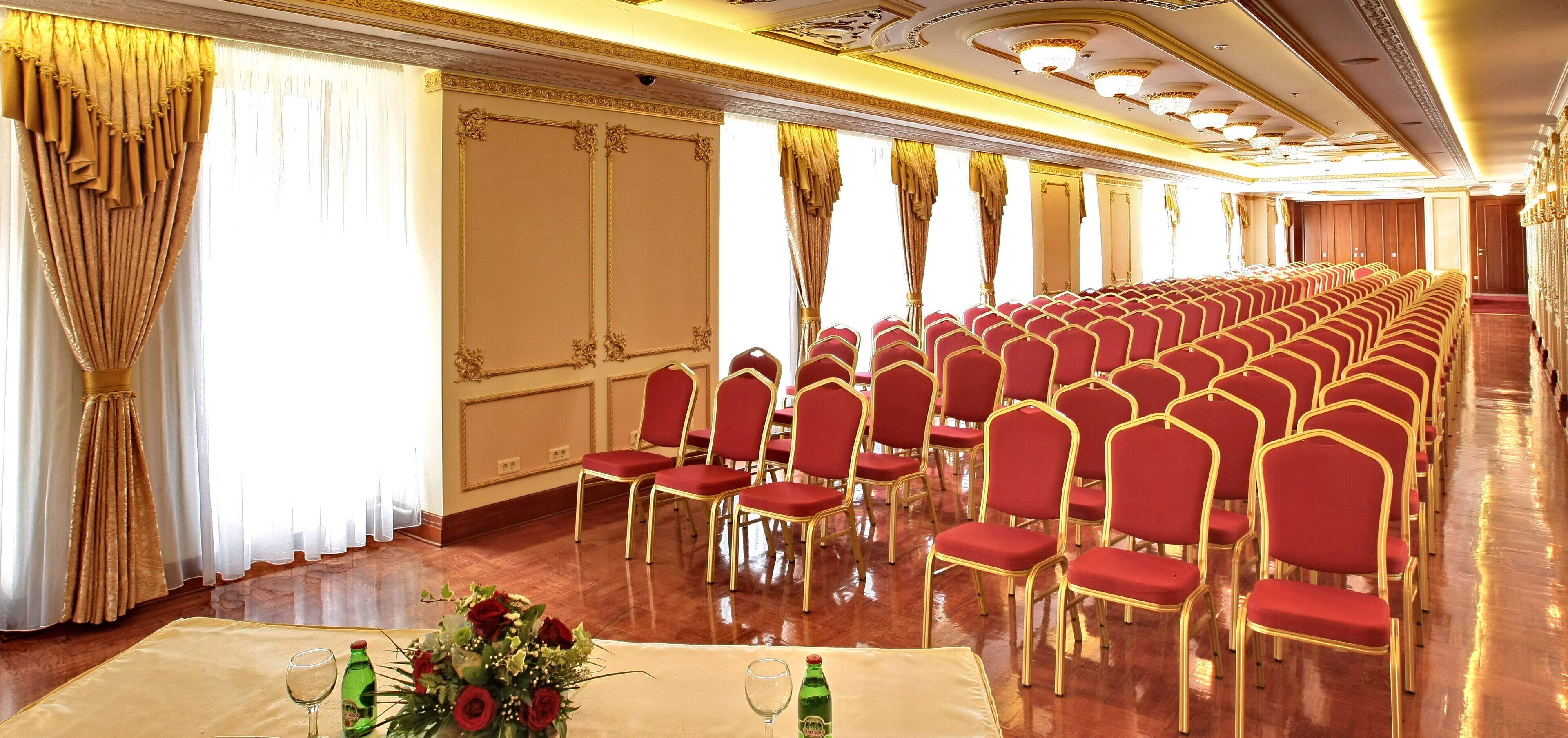 A conference room with rows of red chairs, a polished wooden floor, and a table with a floral arrangement.