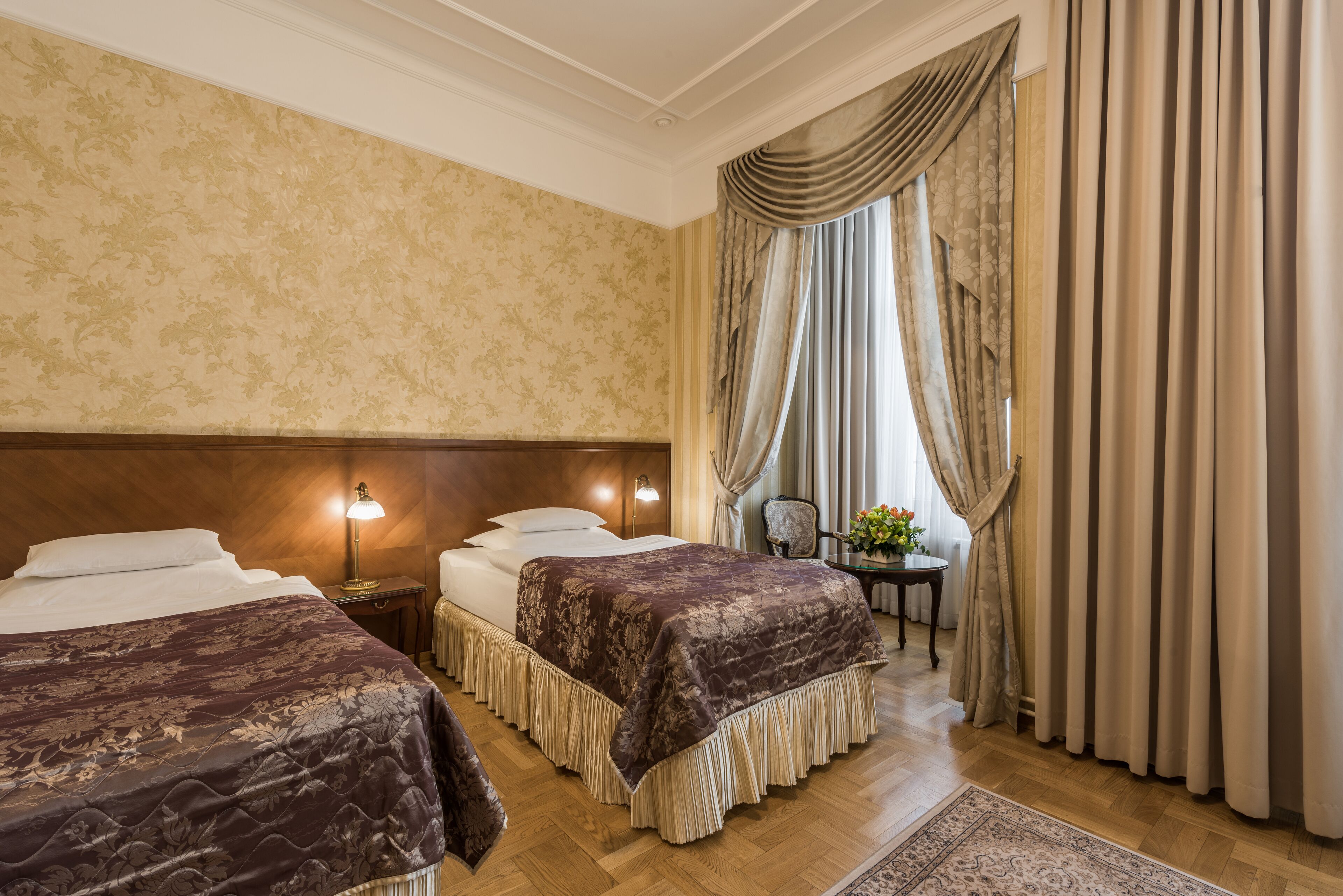 Habitación de hotel con dos camas, cabecera de madera, mesita de noche con lámpara, una pequeña mesa con flores y vistas al exterior a través de cortinas.