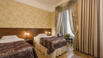 Une chambre d’hôtel avec deux lits, une tête de lit en bois, une table de chevet avec une lampe, une petite table ornée de fleurs et une vue sur l’extérieur à travers des rideaux.