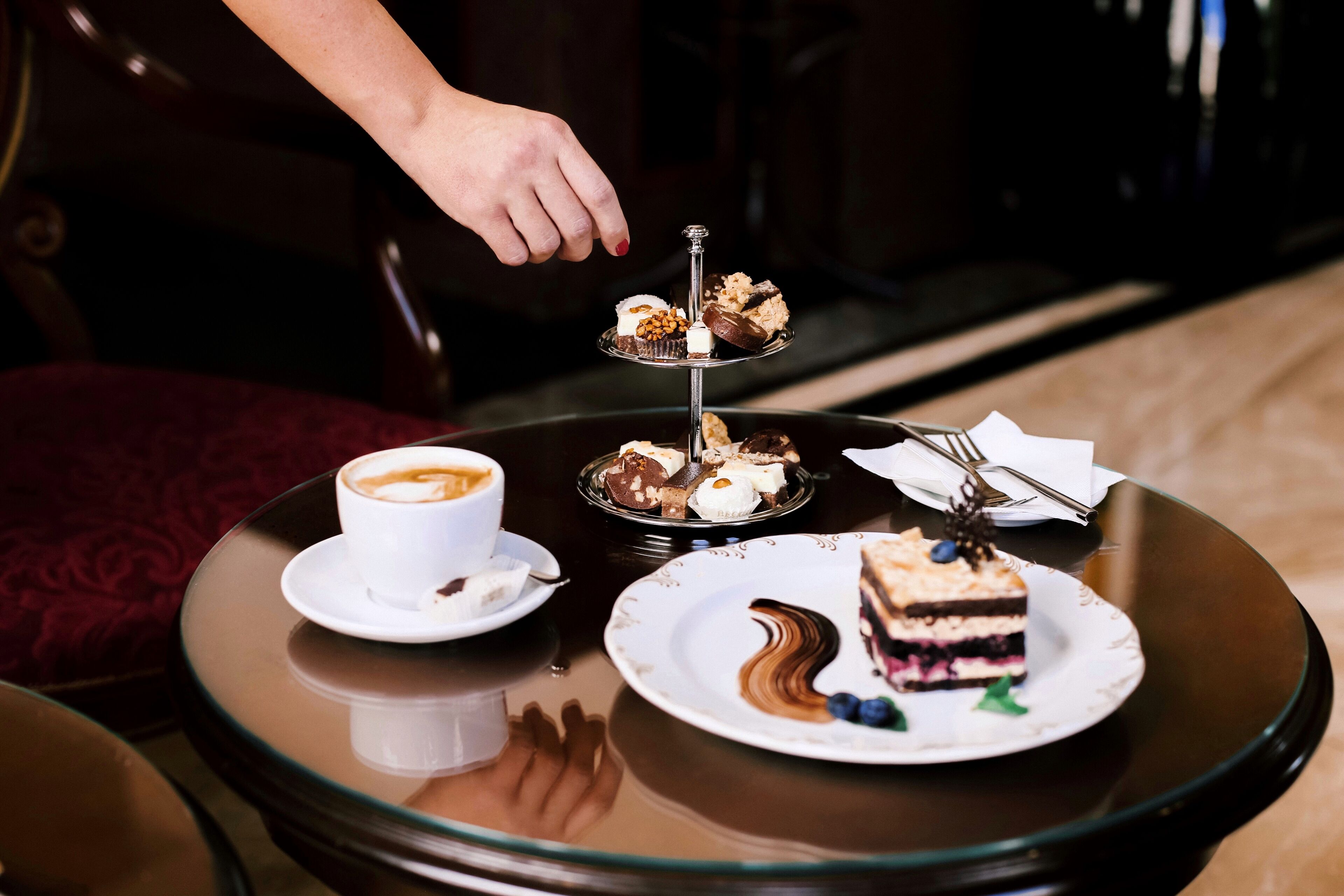 A round glass table with a white cup of coffee, a plate with a slice of cake, and a three-tiered dessert stand with various sweets.