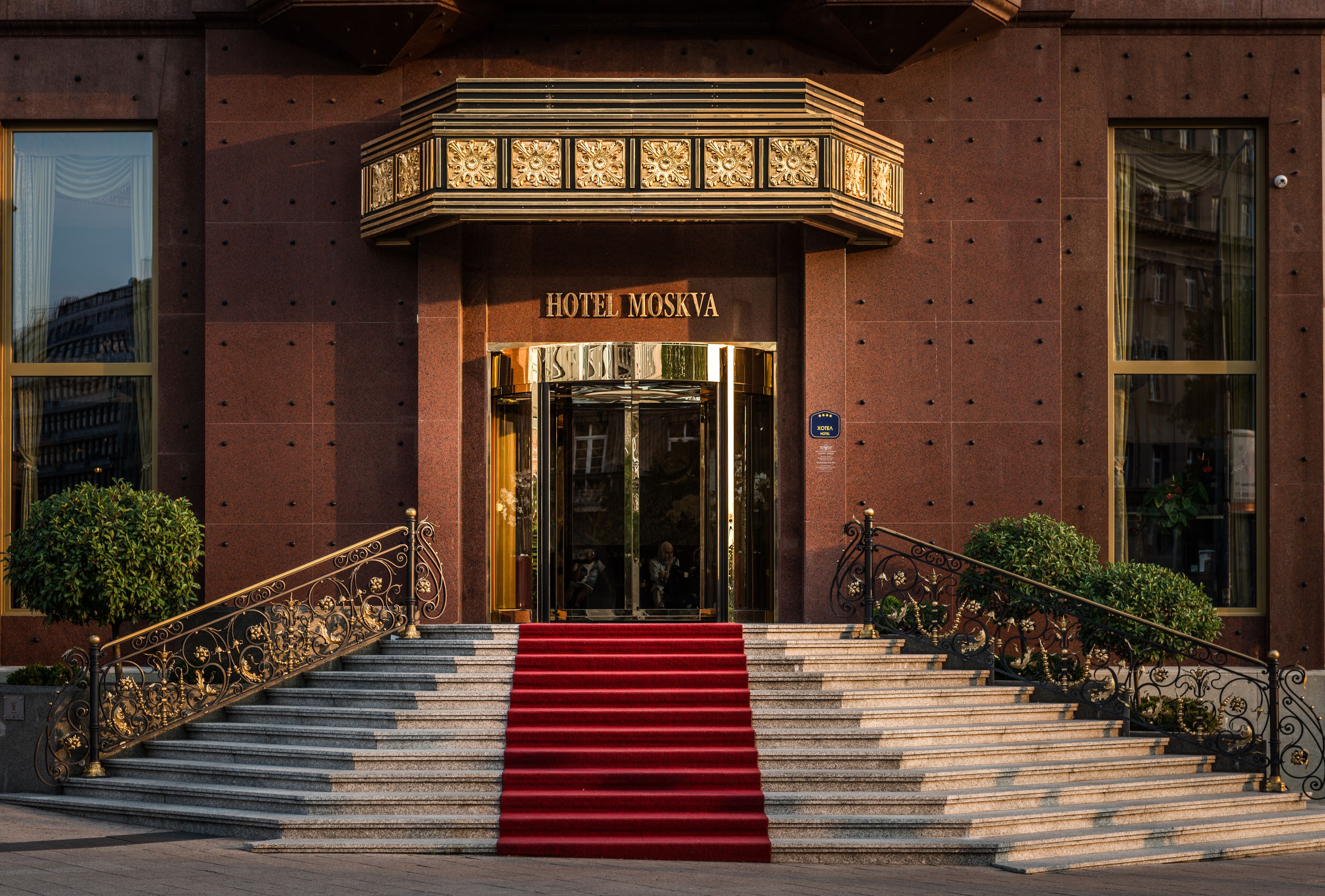 A grand entrance with a red carpet leading to a double door, flanked by ornate railings and a decorative canopy above.