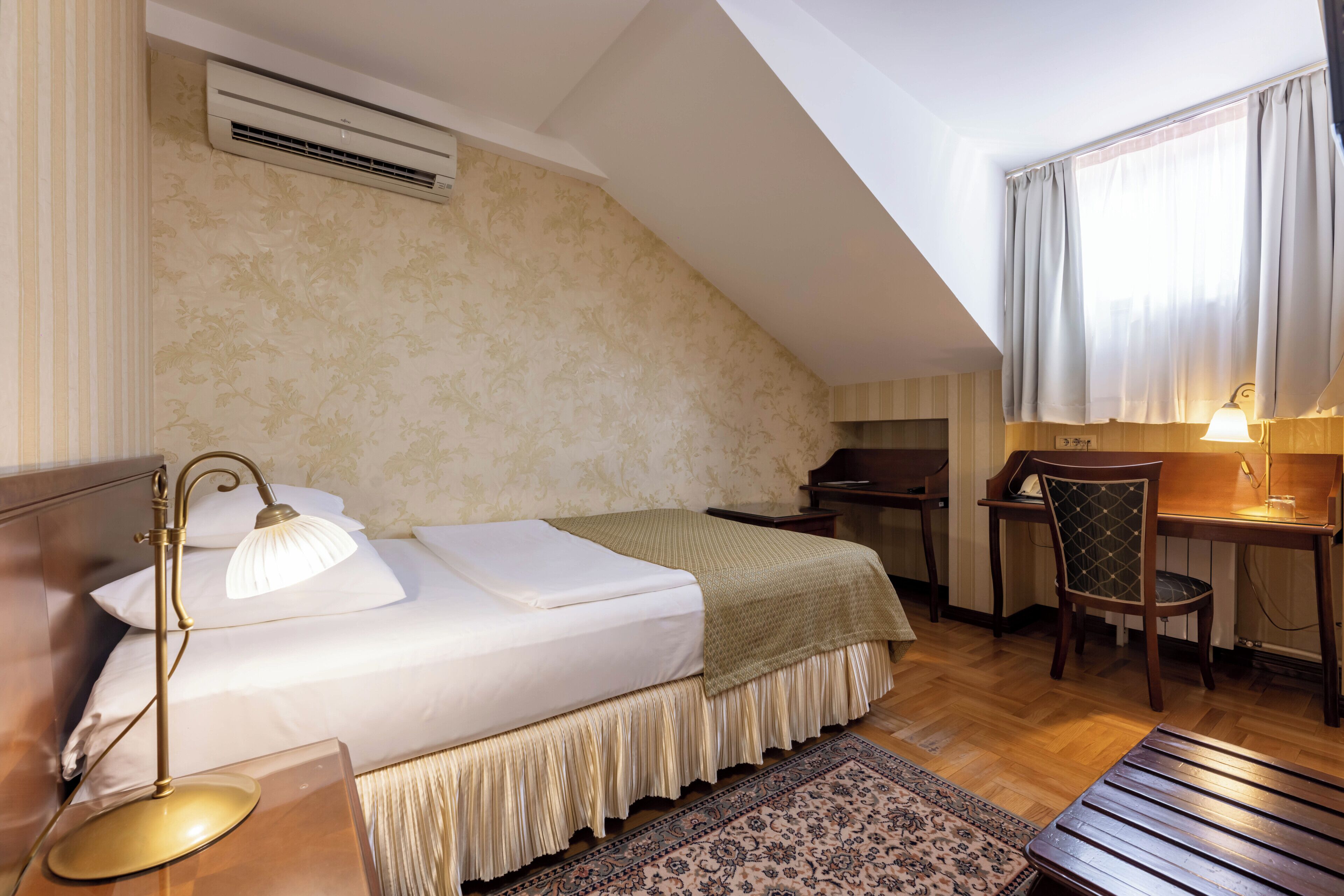 Ein Schlafzimmer mit Bett, Nachttischlampe, Schreibtisch, Stuhl und einem Fenster mit Vorhängen.