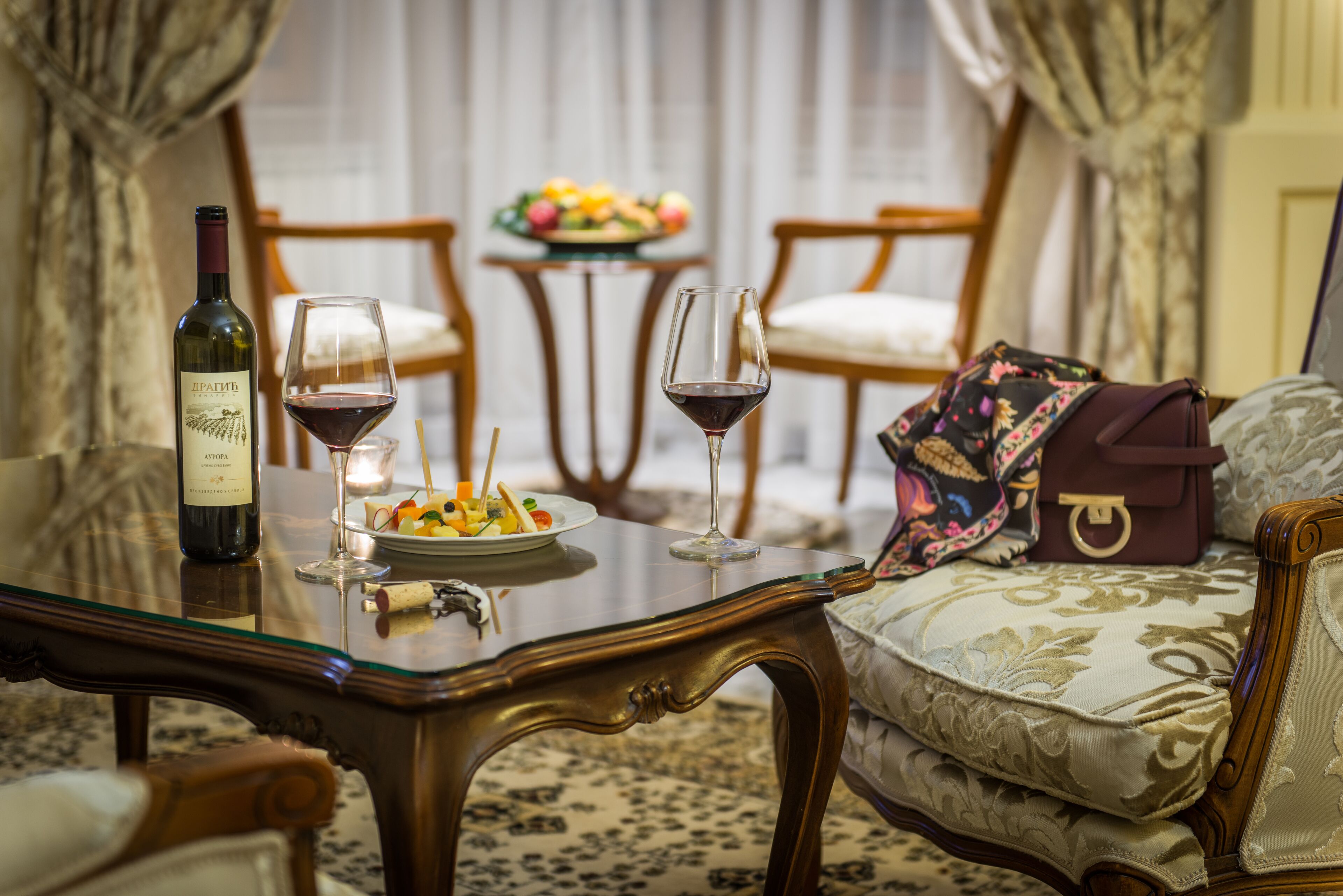 A dining table with wine bottles, glasses, and a fruit plate. There are chairs and a small table with a fruit bowl in the background.
