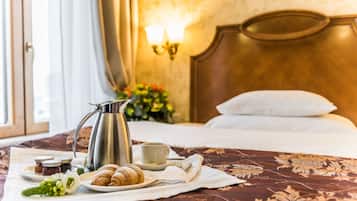 A neatly made bed with a floral bedspread, a bedside table with a coffee pot, a plate of croissants, and a cup of coffee.