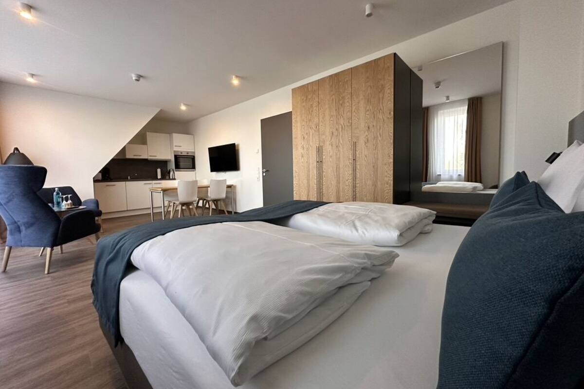 A modern hotel room with a large bed, a desk, and a kitchenette in the background.