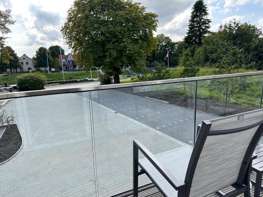 Un área de estar al aire libre con barandilla de vidrio, un sendero empedrado y vistas a una calle residencial con casas y árboles.