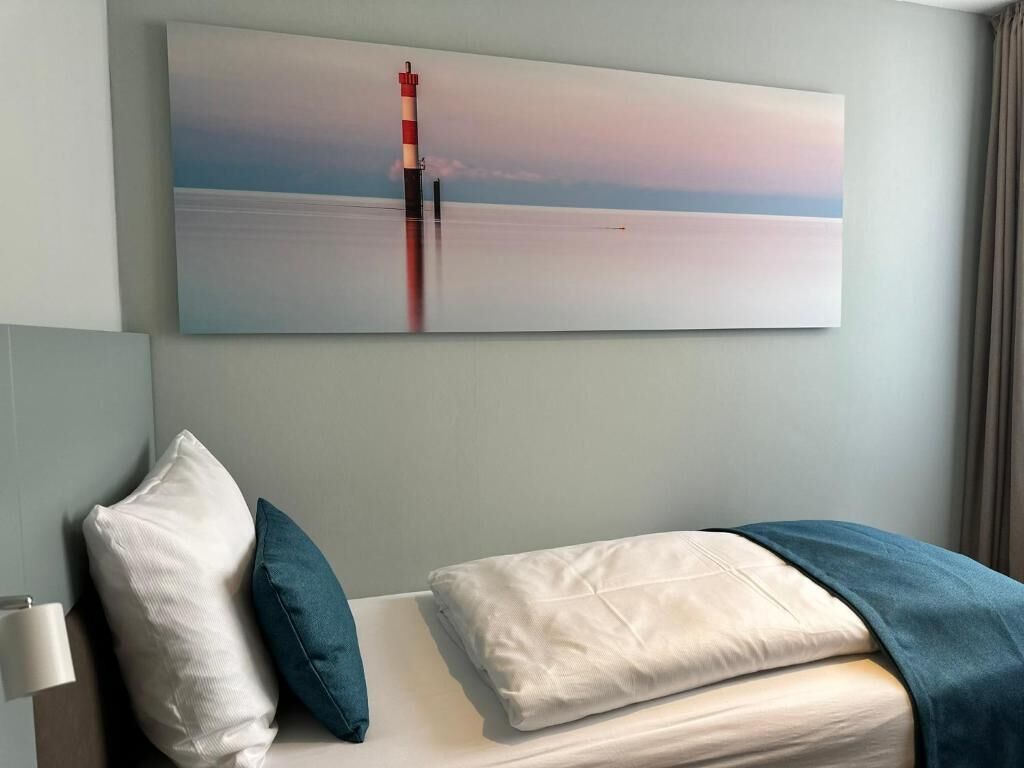 Quarto de hotel com cama, travesseiros, mesa de cabeceira, luminária e uma grande pintura de um farol na parede.