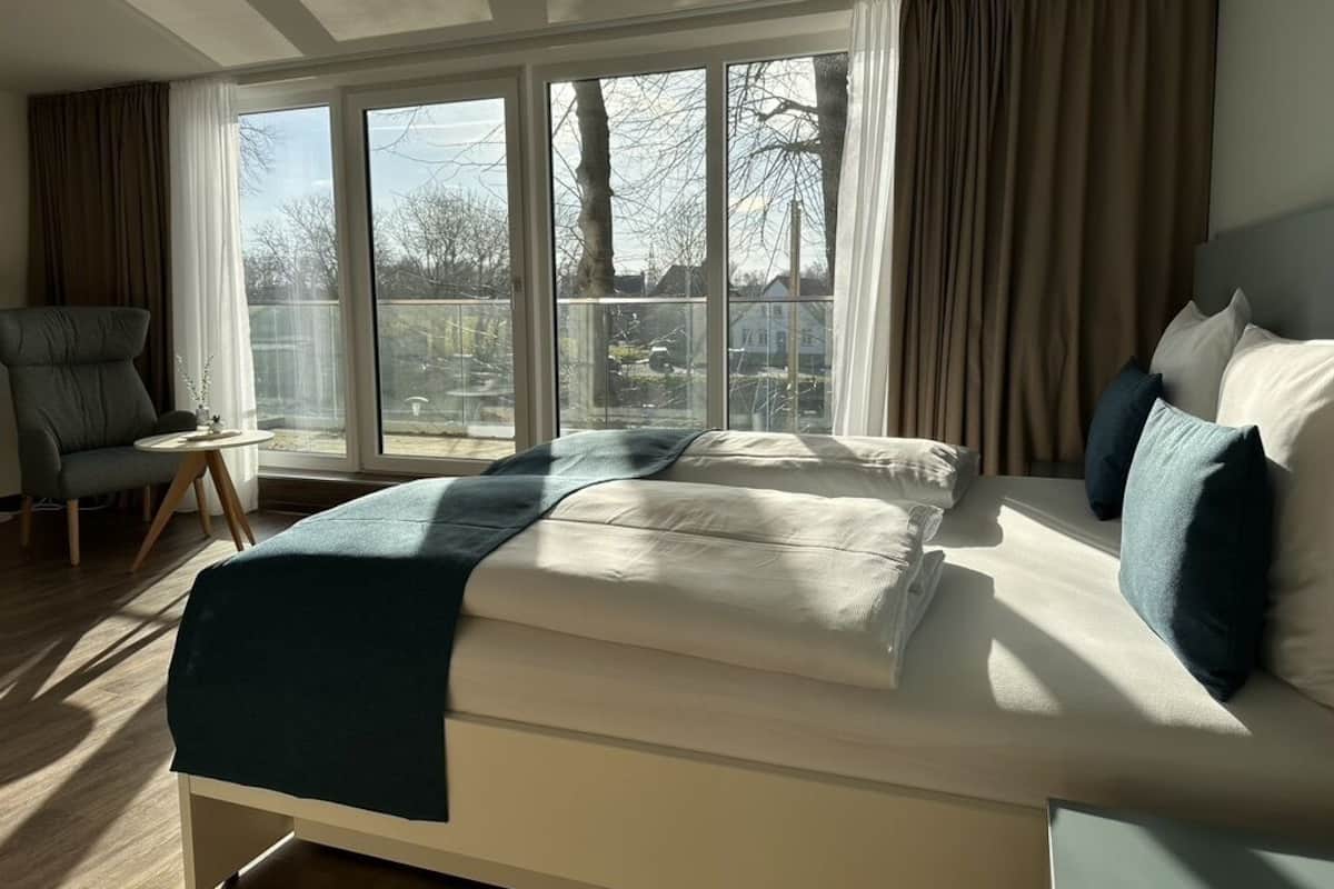 A modern hotel room with a large bed, a chair, and a view of the outdoors through large windows.