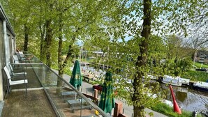 A balcony with glass railings overlooking a river, outdoor seating, and trees. - Hotel Schöne Aussicht, Stilvoll modern, direkt am Rüstersieler Hafen (Wilhelmshaven)