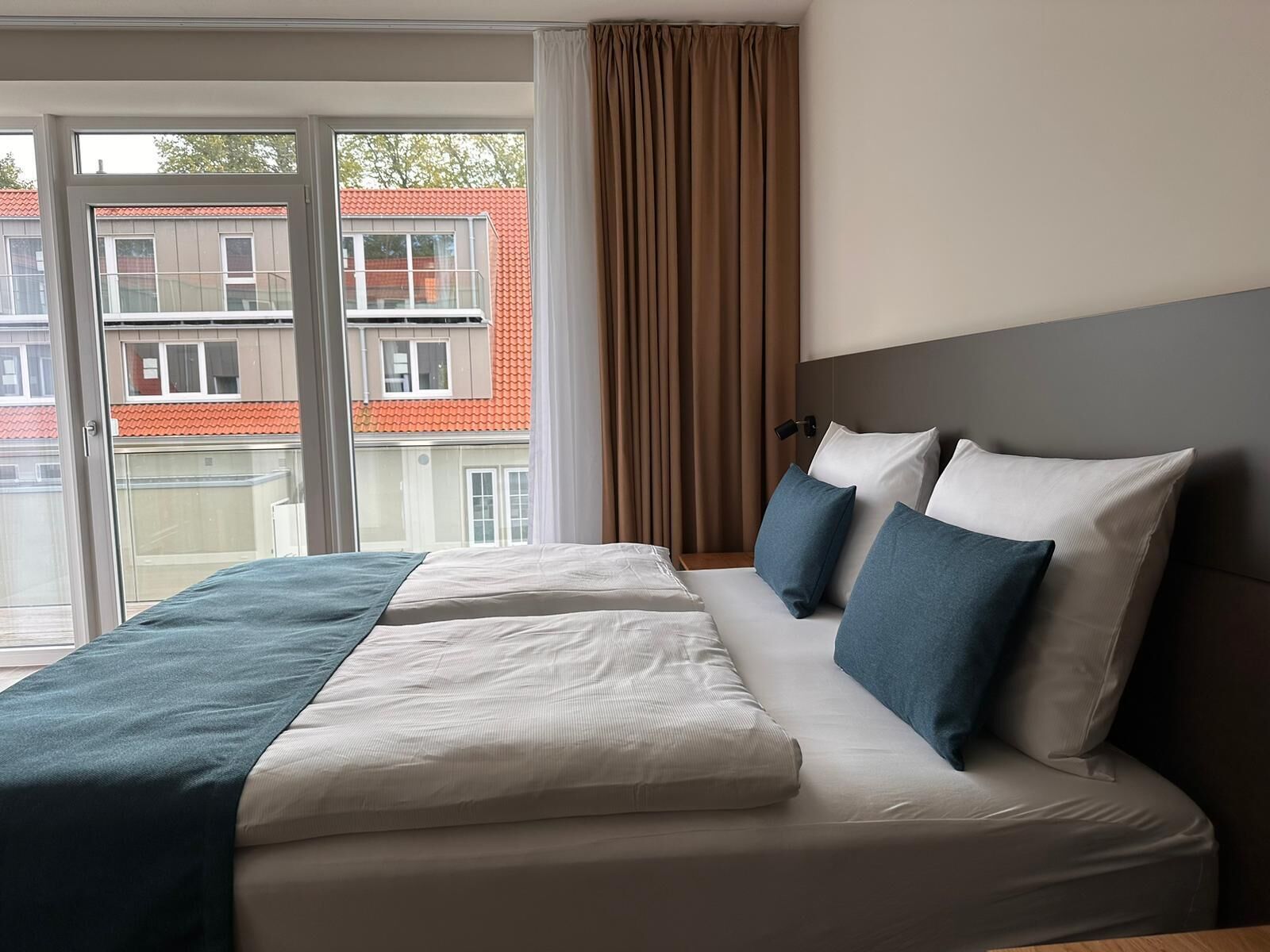 A hotel room with a large bed, a nightstand, a lamp, and a view of buildings through the window.