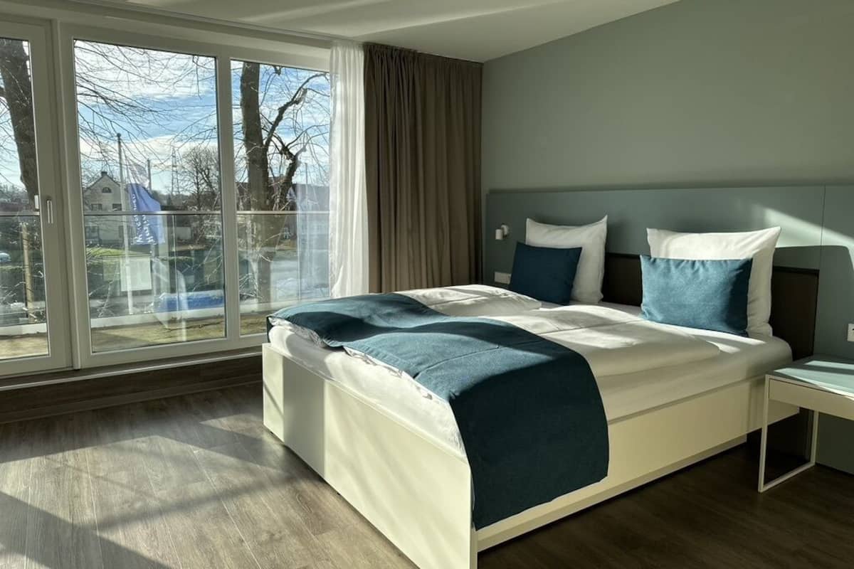 A modern bedroom with a large bed, a nightstand, and a view through a large window.