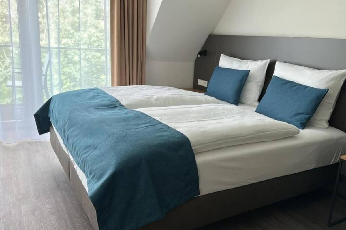 A modern hotel room with a large bed, a bedside table, and a view of greenery through the window.
