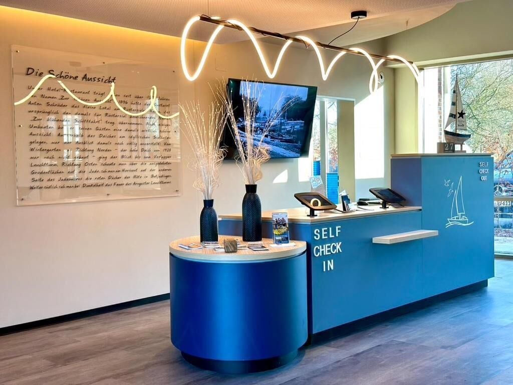 Een receptiebalie met een bordje 'Self Check In', een aan de muur bevestigde televisie en een decoratieve vaas met takken.
