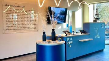 Een receptiebalie met een bordje 'Self Check In', een aan de muur bevestigde televisie en een decoratieve vaas met takken.