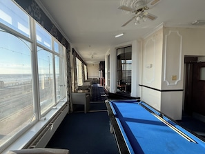Une table de billard dans une pièce dotée de grandes fenêtres, d’un ventilateur de plafond et offrant une vue sur l’océan.