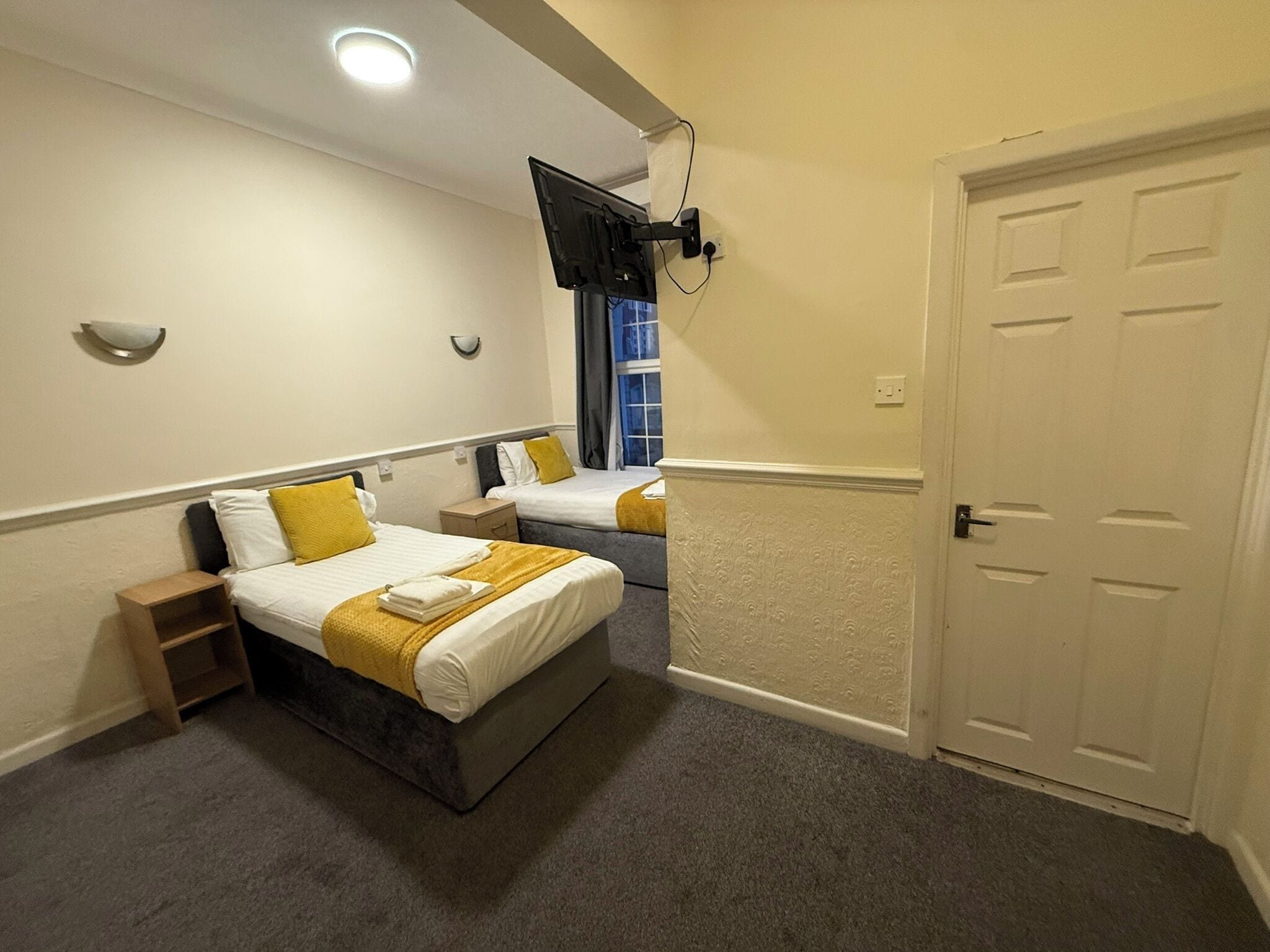 Een hotelkamer met twee bedden, een aan de muur bevestigde televisie, een deur en een raam met gordijnen.