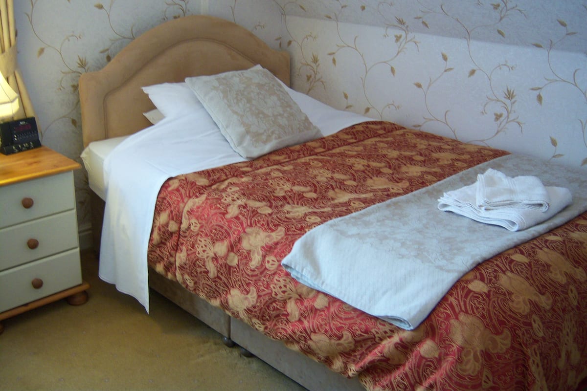 Una cama bien hecha con una colcha estampada con motivos florales, una almohada blanca y una toalla doblada sobre una mesita de noche de madera.