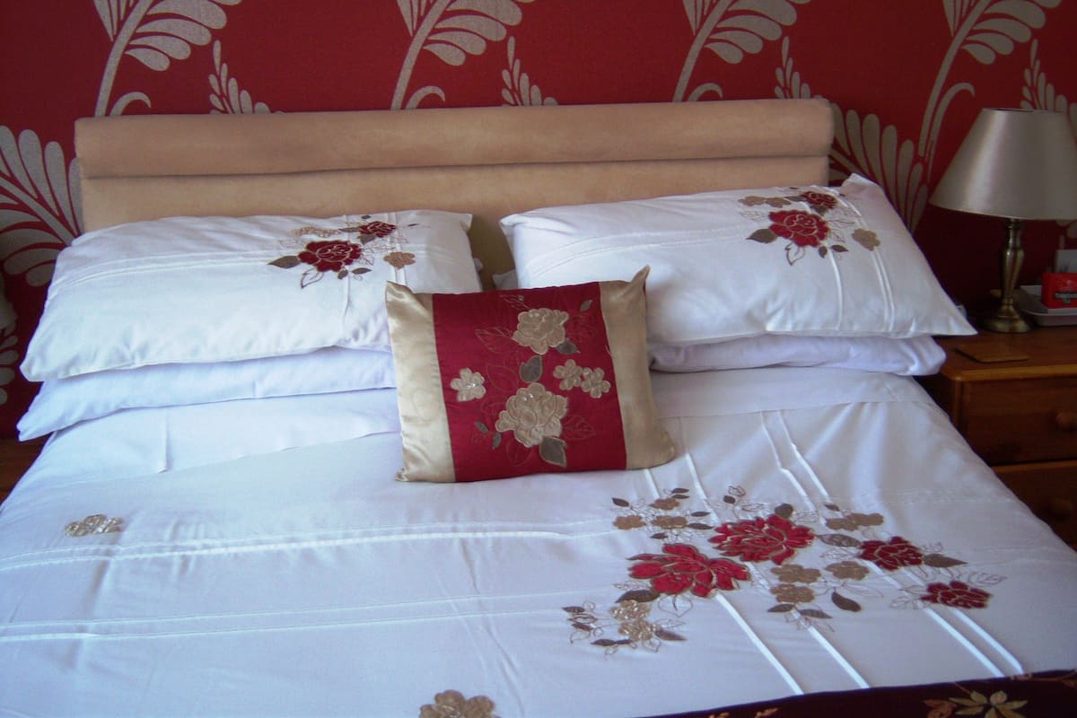 Una cama bien tendida con ropa blanca y almohadas rojas con estampado floral, una mesita de noche de madera con una lámpara y una pared con diseño rojo.