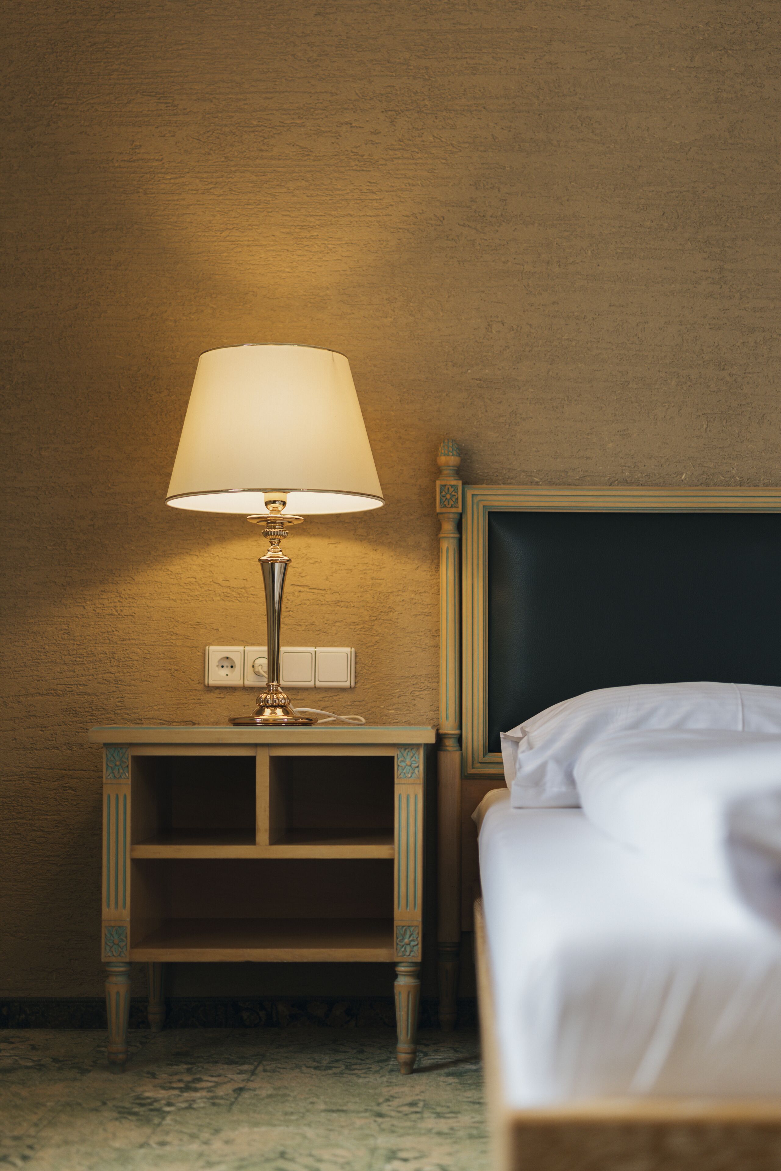 A neatly made bed with a white comforter and pillows, a bedside table with a lamp, and a wall socket.